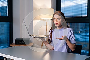 Frustrated young woman confused by laptop computer problem sitting at desk in home office near window evening at late