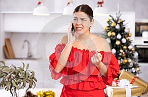 Frustrated young woman in red dress calling on phone next to wrapped gifts and Christmas tree in living room at home
