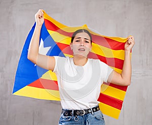 Frustrated young woman waving national flag of Estelada while standing against gray wall