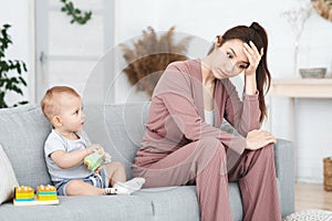 Frustration Of Motherhood. Depressed Young Woman Sitting Next To Her Toddler Baby