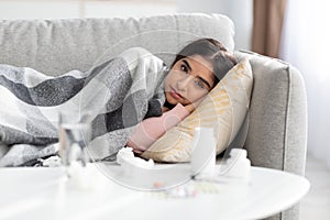 Frustration young hindu lady covered with plaid, suffer from flu, lies on sofa in room interior with water and pills