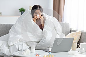 Frustration young indian lady wrapped in blanket blows nose in napkin on sofa with laptop and medicine