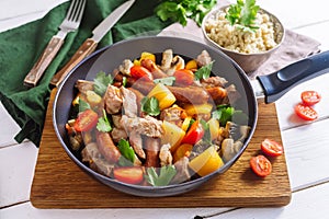 Frying pan with chopped pork meat, yellow bell pepper, cherry tomatoes, sausages, mushrooms, parsley leaves. A frying pan stands o