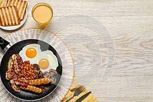 Frying pan with cooked traditional English breakfast and glass of juice on white wooden table, flat lay. Space for text