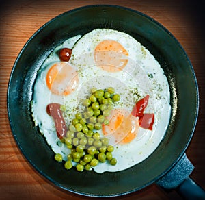A frying pan with three fried eggs as well as peas and ketchup on a wooden background