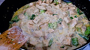 Frying spicy chicken with broccoli in cream on a black pan. Mixing with wooden spatula. Cooking dinner at home on a week night.