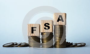 FSA - acronym on wooden cubes. Which stand on stacks of coins on a light background