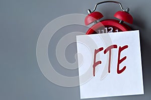 FTE - acronym on a white piece of paper on a gray background. Red alarm clock with white piece of paper