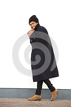 Full body african american male fashion model walking and smiling with winter jacket and beaning by white wall