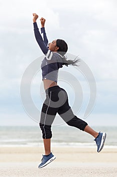 Full body african american woman running and jumping with arms raised at beach