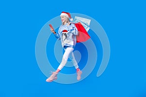 Full body cadre of browsing smartphone young blogging woman run jump air wear red santa hat bring bags isolated on blue