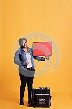 Full body of deliverywoman pointing at red speech bubble advertisting takeaway food service