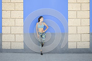 Full body fit Caucasian woman wearing a blue sports bra and leaning against a wall