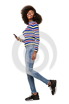 Full body happy young black woman walking with cellphone against isolated white background