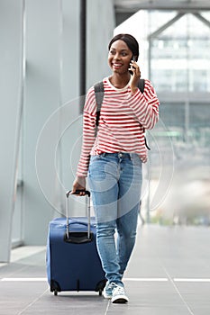 Full body happy young black woman walking with suitcase in airport terminal with cellphone