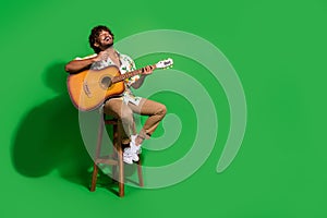 Full body photo of attractive young man sit chair sing play guitar wear trendy citrus print clothes isolated on green