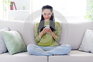 Full body photo of calm peaceful afro american woman sit sofa look hold phone indoors inside house home