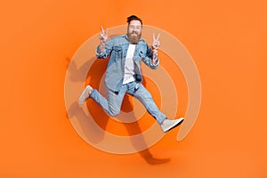 Full body photo of energetic man jumping demonstrate v-sign isolated on orange color background