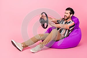 Full body photo of funny aggressive young man lying beanbag with steering wheel beep when see another car isolated on photo