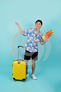 Full body photo of handsome Asian tourist in summer clothes with water gun and luggage during Songkran festival on blue background