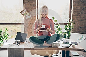 Full body photo of happy positive woman company owner sit on table crossed legs rest relax hold coffee cup drink