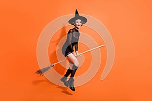 Full body photo of nice young woman straddle wooden broom fly wear stylish black halloween witch look isolated on orange photo