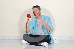 Full body portrait of adult Asian man sitting cross legged looking to his mobile phone with happy expression photo