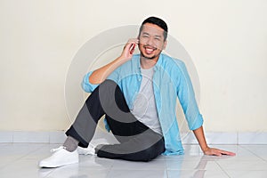 Full body portrait of adult Asian man sitting on the floor smiling friendly while calling someone photo