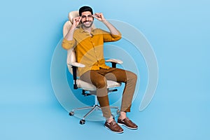 Full body portrait of cheerful young man sit chair hands touch eyeglasses isolated on blue color background