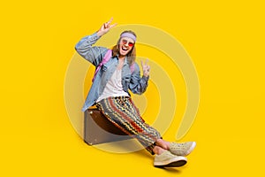 Full body portrait of cool young man sit valise show v-sign wear denim shirt isolated on yellow color background