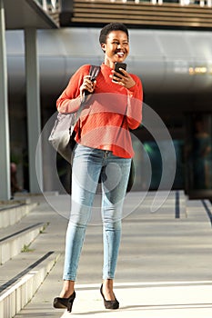 Full body happy young african woman standing outside in city with cellphone and purse