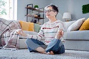 Full body portrait of nice positive aged lady sit carpet floor meditate daylight modern apartment inside