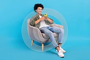 Full body portrait of positive young man sit chair use smart phone isolated on blue color background