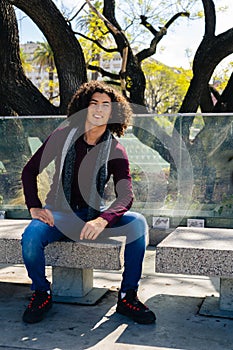 Full body portrait of a smiling young curly haired Latino man sitting on a concrete bench in the shade of an old tree. Vertical