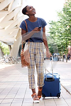 Full body young african american woman walking in city with suitcase and mobile phone