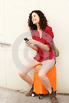 Full body young woman sitting on suitcase and holding mobile phone