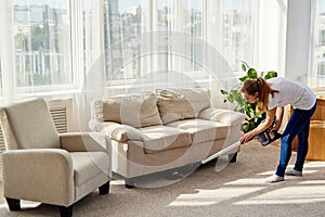 Full body portrait of young woman in white shirt and jeans cleaning carpet with vacuum cleaner in living room, copy space.