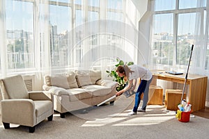 Full body portrait of young woman in white shirt and jeans cleaning carpet with vacuum cleaner in living room, copy space.