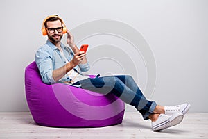 Full body profile photo of funny guy sitting cozy armchair holding telephone using cool earflaps listen youth song wear