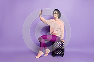 Full body profile photo of pretty lady waiting flight hold telephone sitting on bag making selfies wear hat sun specs