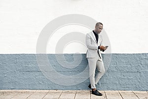 Full body serious african american businessman leaning against wall by street with cellphone