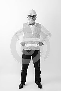 Full body shot of handsome senior bearded man construction worker standing with hands on hips while wearing eyeglasses