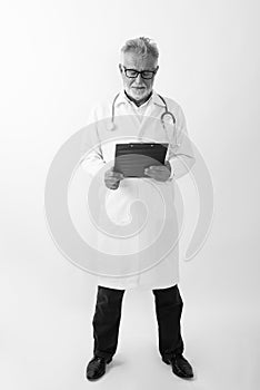 Full body shot of handsome senior bearded man doctor standing while reading on clipboard against white background