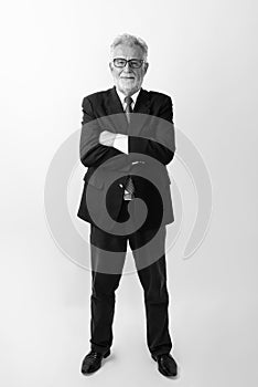 Full body shot of happy senior bearded businessman smiling and standing while wearing eyeglasses with arms crossed