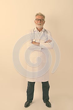 Full body shot of happy senior bearded man doctor smiling while standing with arms crossed against white background