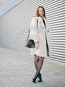 Full body shot of young trendy dressed serious caucasian woman posing outdoor on urban city blurred geometric background cold