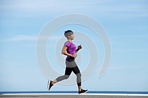 Full body side fit young african woman running outdoors against blue sky