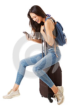 Full body side of happy asian female traveler sitting on suitcase and looking at cellphone against isolated white background