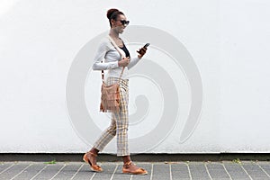 Side portrait young african american woman walking against white wall with cellphone