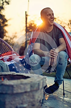 Full body vertical shot of cheerful man with the flag of USA on his back, relaxing in a cozy chair outdoors